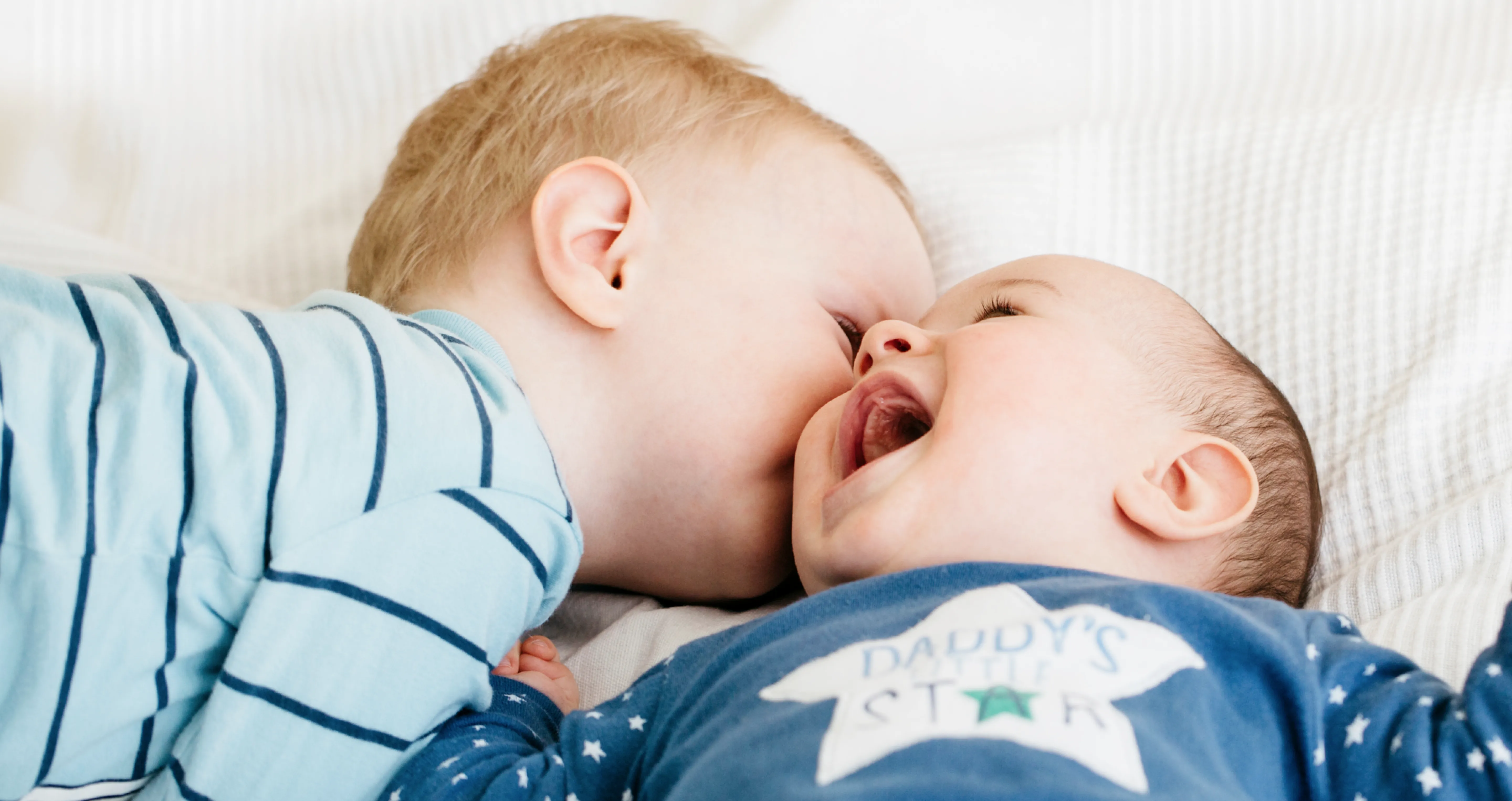 Two babies lying on a bed, laughing and playing, with one baby gently biting the other's cheek in a playful moment.