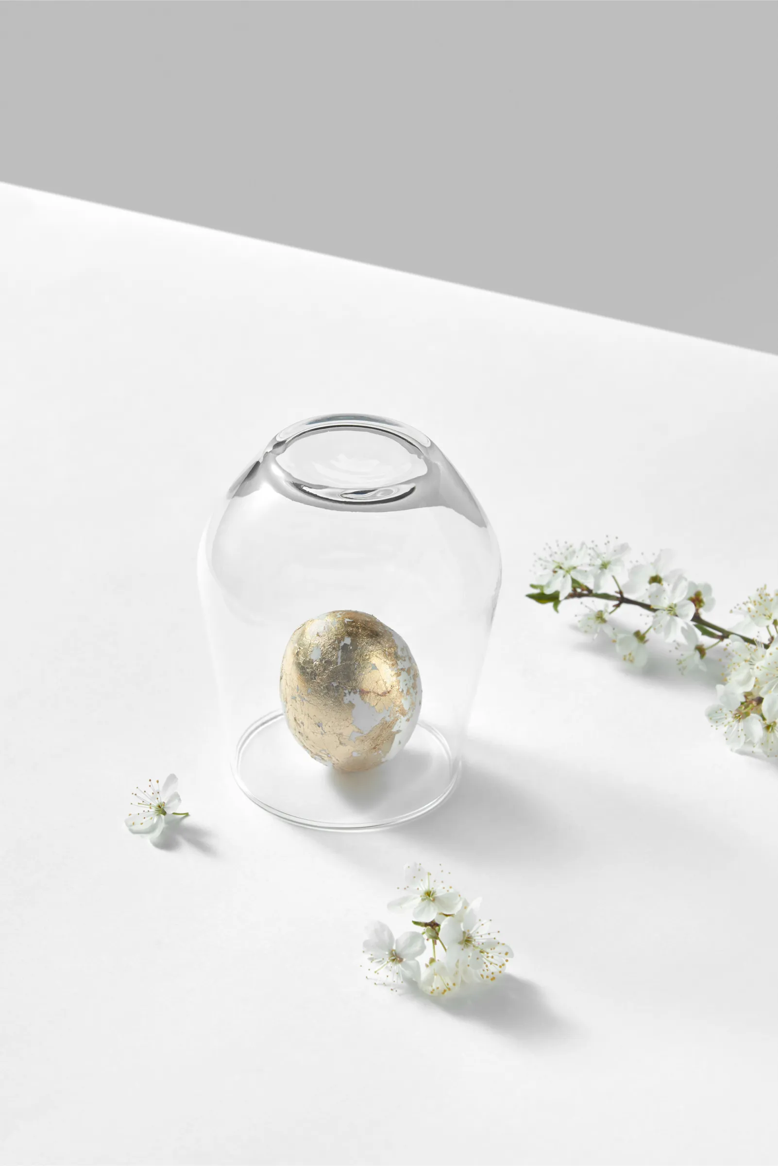A golden speckled egg displayed under a clear glass cloche, accompanied by delicate white blossoms on a clean, white surface with a soft gray background.
