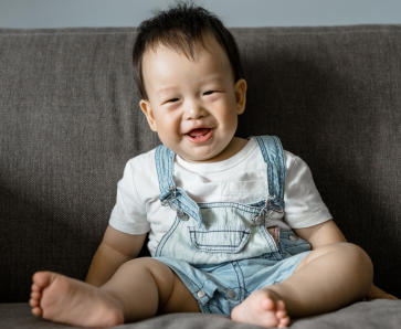 A happy baby with a big smile, sitting on a gray couch in a light blue denim jumpsuit, exuding joy and playfulness.